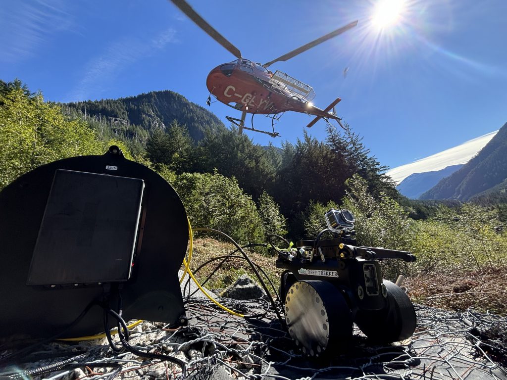 Helicopter supporting remote subsea inspection equipment deployment in a mountainous coastal environment.